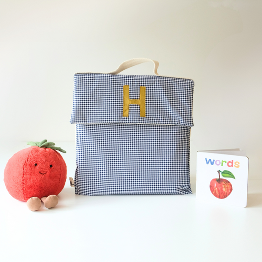 A blue and white checkered kids' bag with a personalized 'H' on it, displayed next to a red plush toy and a small booklet with the text 'WORDS'.