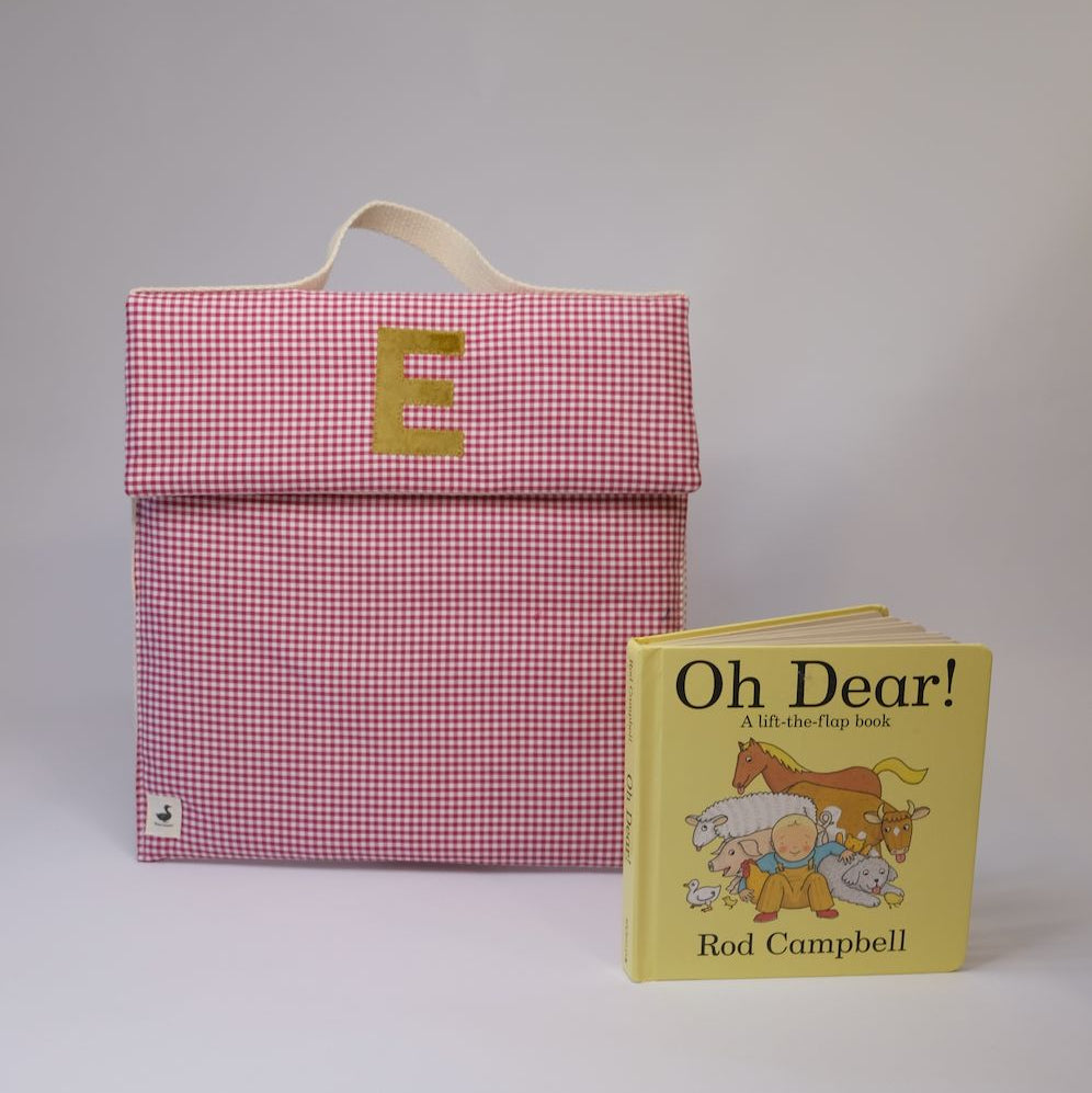Pink checkered bag with a gold letter 'E' next to a yellow book titled 'Oh Dear!' by Rod Campbell on a light gray background.